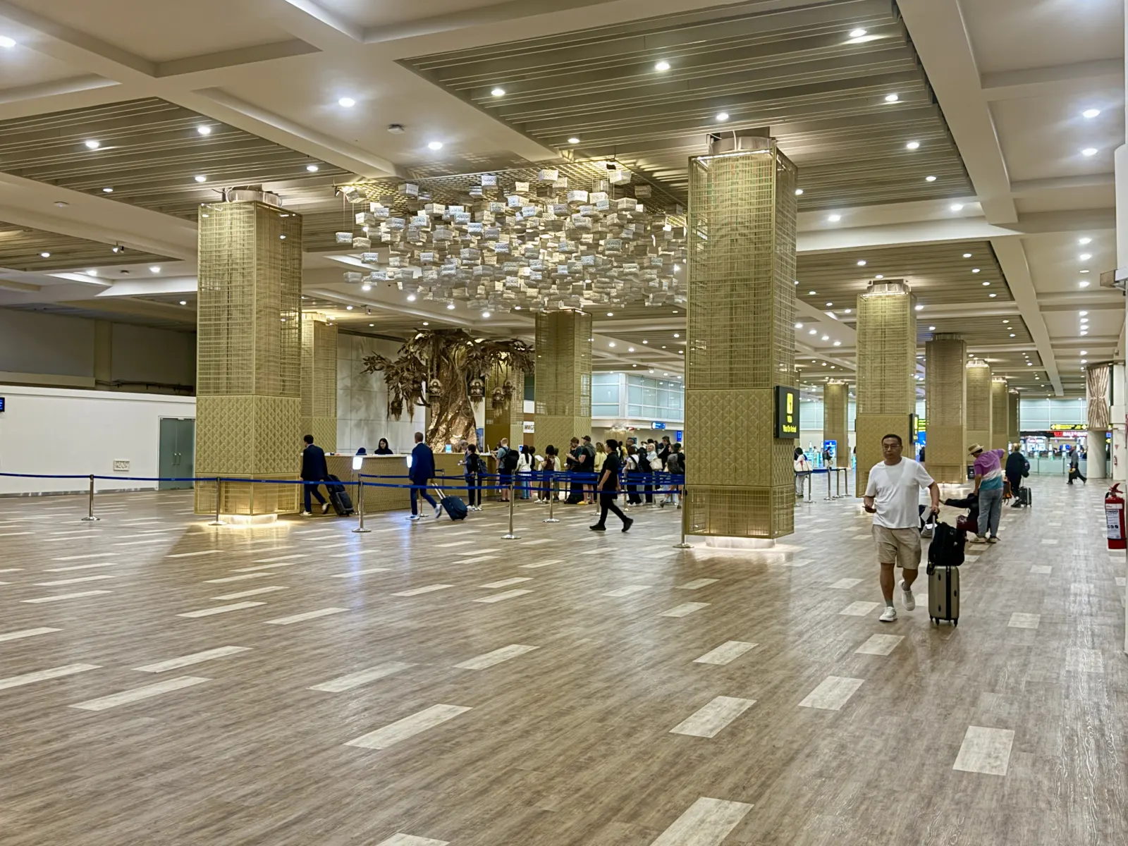 Spacious hall at Bali's international terminal with gold-clad pillars, a large chandelier and passengers with suitcases.