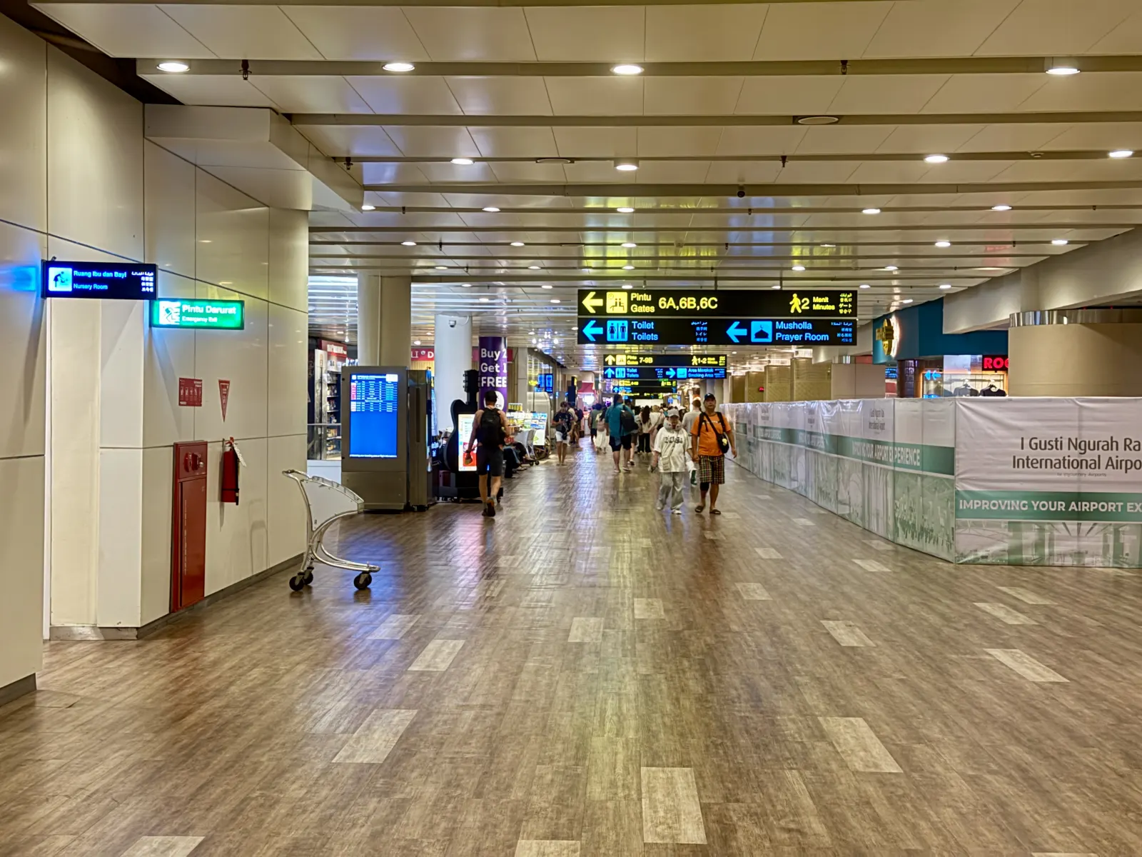 Wide corridor inside the international terminal of I Gusti Ngurah Rai Airport, with overhead direction signs and travellers walking toward the gates.