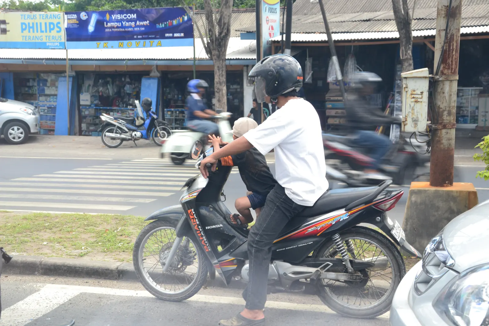 A man in a helmet rides a motorbike with a child seated in front of him past roadside shops in Bali, with other riders behind.